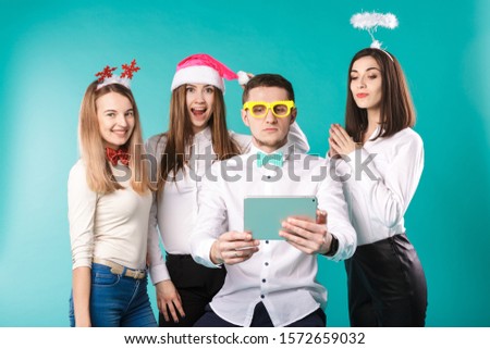 New Year theme Christmas winter office company employees. Group 4 young Caucasian people business smile holiday funny hats accessories glasses take photo yourself selfie tablet blued background.