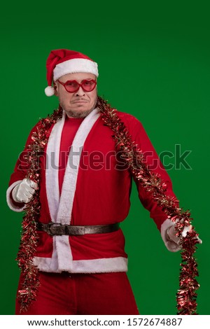 Male actor in a costume of Santa Claus in large pink glasses and with a red garland of tinsel in his hands dancing and posing on a green chrome background