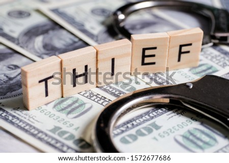 Word made of wooden letters - thief, on a gray background with us dollars next to a magnifier. Crime designation concept. Close up.