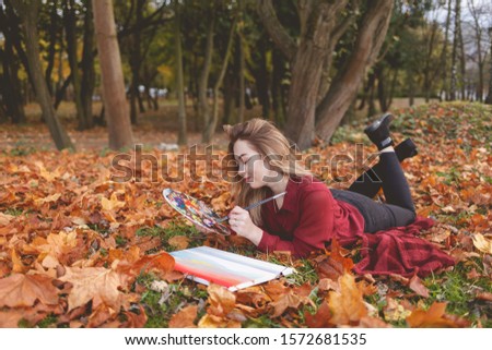 Young woman artist is relaxing in the park for her favorite hobby. Girl draws a picture, holding a palette in hand