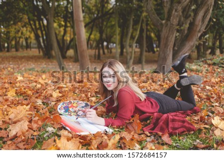 Young artist lies in autumn park on a plaid and paints a picture. 
Girl draws a picture in the park