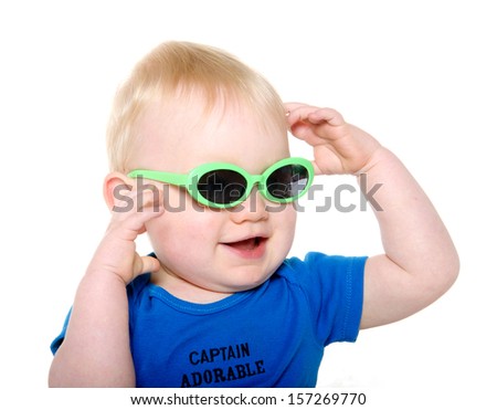Cute 18-month-old baby boy with blond hair and blue shirt wearing green sunglasses on white background