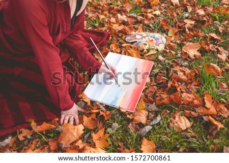 Young girl artist draws a picture kneeling in a park on the grass. 
Girl takes inspiration for creativity sitting in autumn park and painting