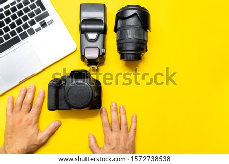 Photo camera and other accessories such as flash light and lens are on the table near laptop on yellow background. Man photographer prepairs photo for editing. View from the top, flat lay.