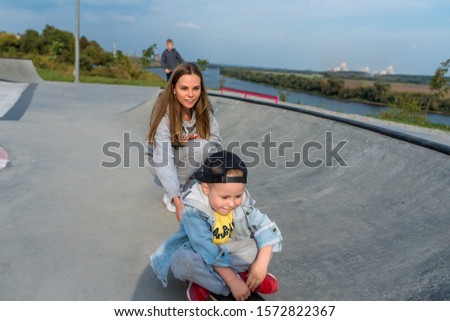 Young mother teaches ride skateboard, son of little boy, summer day. Autumnal clothes, casual clothes. Help and support parenting. Caring for children. Workout balance
