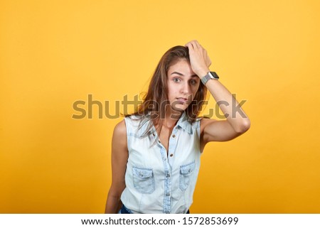 Confused caucasian young woman in blue denim shirt keeping hand on head, looking sad isolated on orange background in studio. People sincere emotions, lifestyle concept.