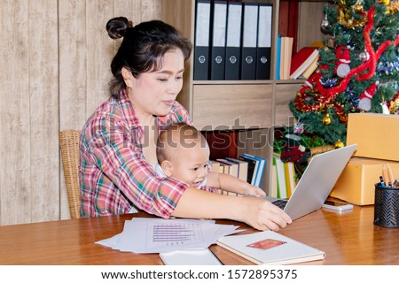 Beautiful Asian mother woman with baby working at home using laptop, smartphone checking package products, small business startup entrepreneur, freelance lifestyle shopping online holiday season
