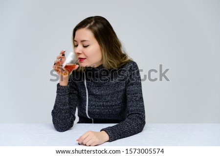 Portrait of a sitting woman at the table talking with curly hair with emotions in a gray dress on a white background with a glass of whiskey in her hands. Female alcoholism concept.