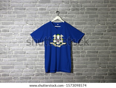Connecticut flag on shirt and hanging on the wall with brick pattern, Three bunches of grapes on royal blue color. text below "Qui Transtulit Sustinet", Latin for "He who transplanted sustains"