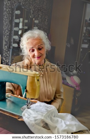 beautiful long grey haired senior woman sews on sewing machine. Dressmaker work on the sewing machine. Tailor making a garment in her workplace. Hobby sewing as a small business concept