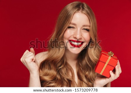 Image of young happy smiling woman posing isolated over red wall background with bright red lipstick holding christmas present gift.
