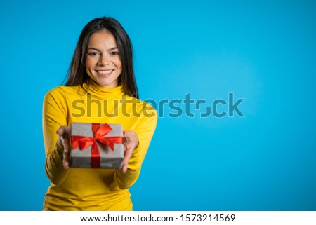 Pretty brunette woman gives gift and hands it to the camera. She is happy, smiling. Girl on blue background. Positive holiday picture