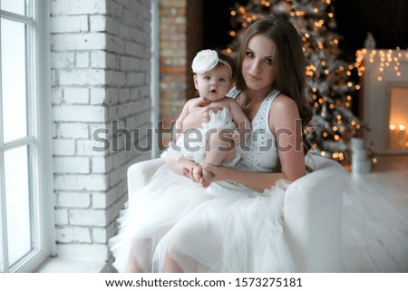 Portrait of happy mother and adorable baby celebrate Christmas. New Year's holidays. Toddler with mom in the festively decorated room with Christmas tree and decorations. Mother playing with baby girl