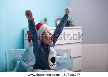 Funny child boy in Santa Clause hat and pajamas waking up in Christmas morning. Happy child sleep in white bed in child bedroom at home. Happy Xmas and New Year holiday! Christmas gift. 