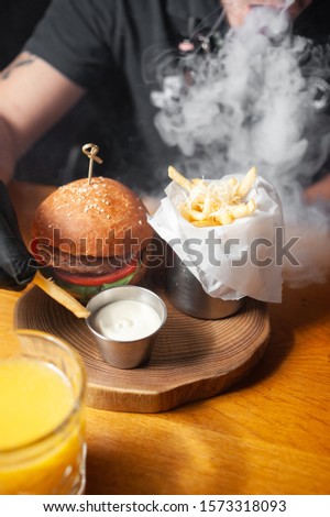 Burger on a wooden board with french fries and sauce in smoke from a hookah
