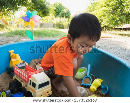 Asian child boy in orange t-shirt playing toy and sand in sandbox with happy face outdoor with rural natural background. Family relax and freedom time in summer holiday.