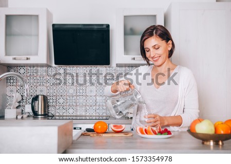 Young sincerely smiling female pouring a purified mineral water into transparent glass decanter at modern kitchen. Plenty of apples, grapefruits, kaki and oranges fruits, berries are on the tables.  Royalty-Free Stock Photo #1573354978