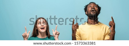shocked interracial couple pointing with fingers up on blue background, panoramic shot
