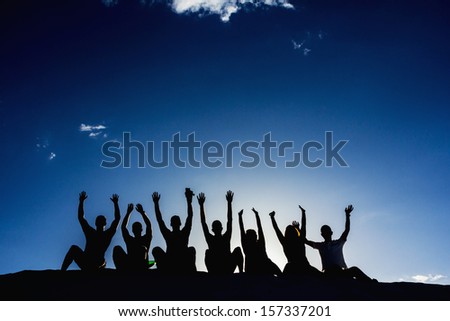 Silhouettes of seven young people rising their hands on a blue clear sky