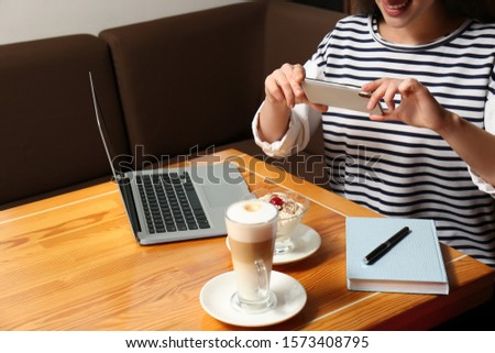 Young blogger taking picture of dessert and latte at table in cafe, closeup