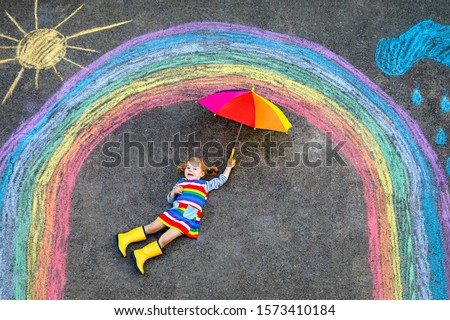 Happy little toddler girl in rubber boots with rainbow sun and clouds with rain painted with colorful chalks on ground or asphalt in summer. Cute child with umbrella having fun. creative leisure