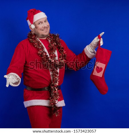Emotional male actor in a costume of Santa Claus holds a Christmas sock in his hands with gifts and poses on a blue background