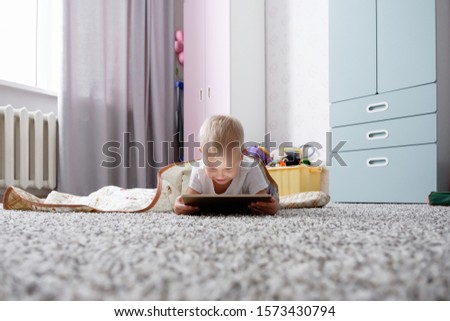 Little positive kid boy watching cartoons on tablet while lying on the floor in his cozy beautiful children's room. Concept of addiction to gadgets for children and developing online games
