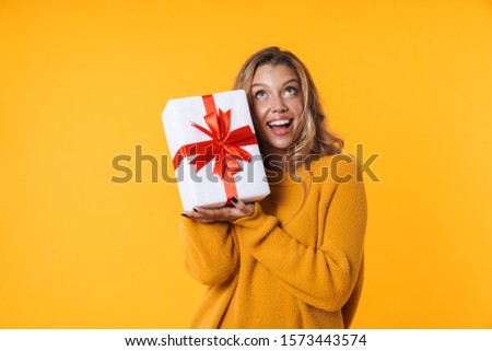 Image of pleased blonde woman in warm sweater smiling and holding present box isolated over yellow background