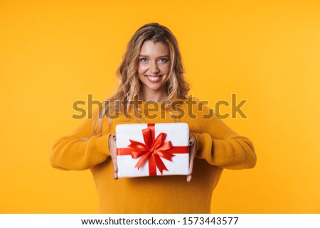 Image of beautiful blonde woman in warm sweater smiling and holding present box isolated over yellow background