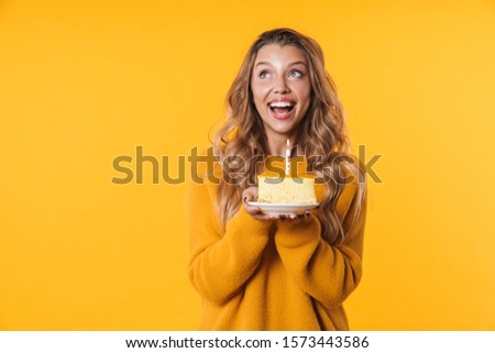 Image of happy blonde woman in warm sweater smiling and holding birthday cake with candle isolated over yellow background