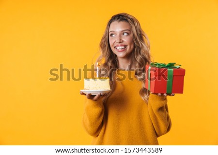 Image of charming blonde woman in warm sweater holding birthday cake and present box isolated over yellow background
