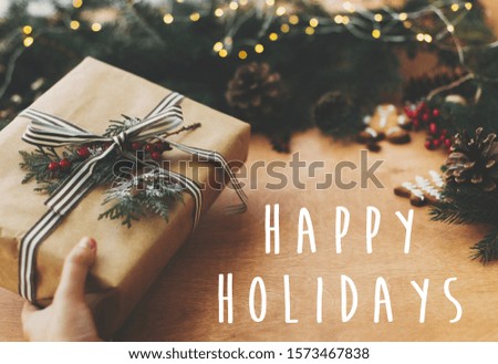 Happy Holidays text sign on stylish rustic christmas gift, pine branches, cones, gingerbread cookies, cinnamon, anise on rural wooden table, Season's greeting card