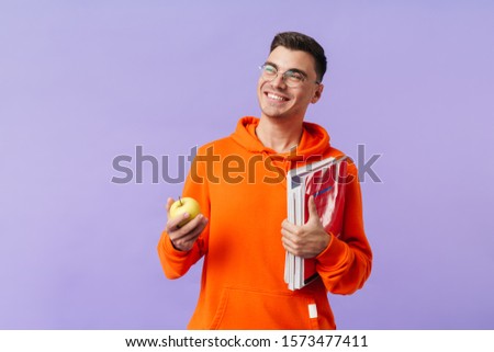 Photo of positive happy young man student posing isolated over purple background holding copybooks and apple.