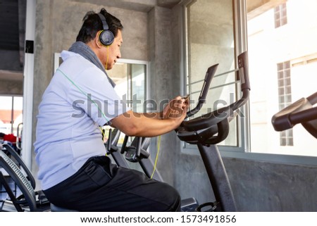 Senior Asian man in sportswear listen to music and training biking cardio at fitness gym.