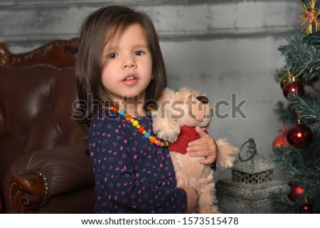 little dark-haired girl sitting with a bear by the Christmas tree at Christmas, portrait