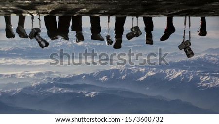 Conceptual scene with photographers sitting on edge of rock with cameras hanging on straps. Silhouettes of legs and cameras against beautiful snow capped mountains. Concept of photography and travel