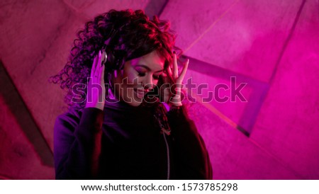 Young beautiful curly woman in big headphones smiles, listens to music, sings along, dances, jumps, closing her eyes, against the background of a wall, neon color, magenta.