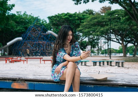 One Asian female in floral dress holding mobile phone in a playground park. She is taking selfie, or could be on video call with family or friends.