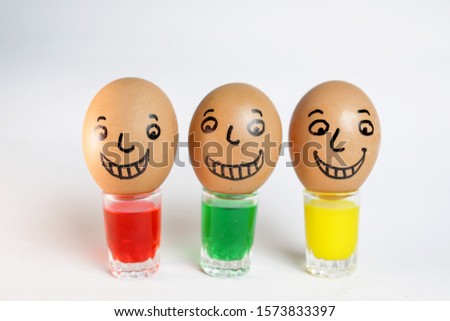 A triad of chicken eggs with paintings of facial characters with happy expressions. Egg on a glass containing colorful water.