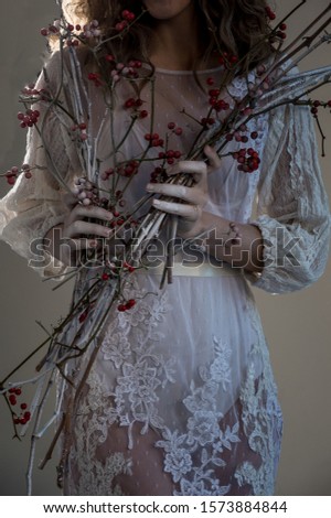 Fragment of a woman in a white dress with snow-covered hands and a branch with red berries in her hands. Winter fairy tale.