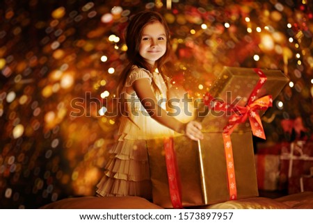 Cute little girl opens the gift box next to a beautiful Christmas tree. Christmas and New Year concept. The atmosphere of magic, lights and sparkle is around.