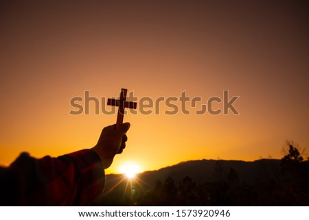 Human hand holding christian cross with a sunset sky background. Christian silhouette concept.
