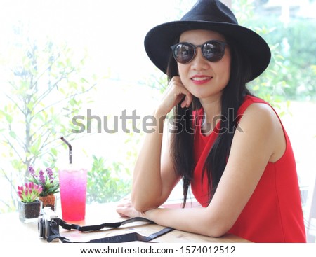 Beautiful Asian young tourist woman wearing hat , sunglasses and red sleeveless sitting with her chin on hand  by the window in coffee shop with camera and red drink on table. 