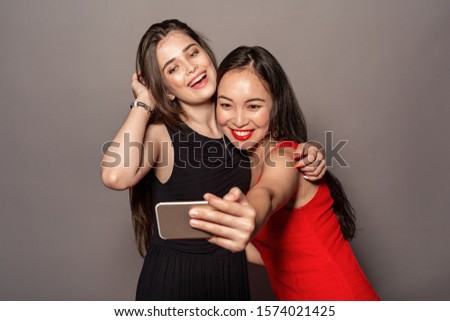 Young women wearing evening dress studio standing hugging isolated on gray background holding smartphone taking selfie photo posing to camera smiling happy