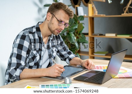 Side view of smart and qualified young adult man using graphic tablet and stylus pencil at home, working on the computer and editing photos
