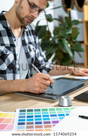Vertical photo of concentrated young adult worker using graphic tablet and stylus pencil in workplace, sitting behind table with color swatches and editing artwork