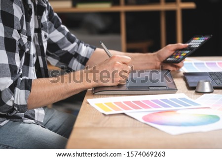 Cropped view of young adult freelancer using color swatches and graphic tablet with stylus, sitting in office, working on computer