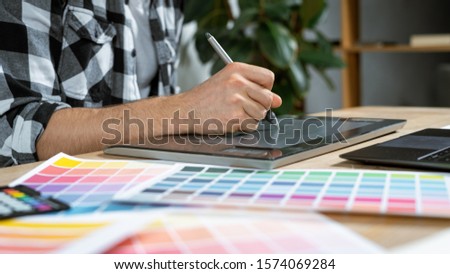 Cropped and panoramic view of young adult illustrator using graphic tablet and stylus pencil, drawing sketch, working at office, sitting behind table with color palette