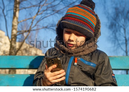 A happy, joyful boy with a smartphone sits on a bench in winter.  The boy listens to his favorite music on the phone. Winter.