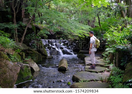 An asian male tourist posing for a picture with green Japanese maple trees and a waterfall in Tokugawa garden, Nagoya, Japan.